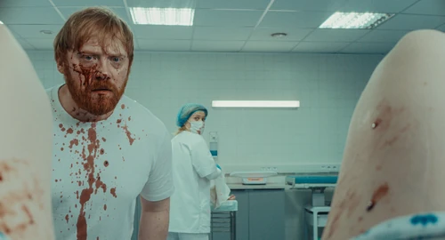 Nightborn: «Un hombre con el rostro y la camiseta cubiertos de sangre observa algo fuera de plano en una sala médica.»