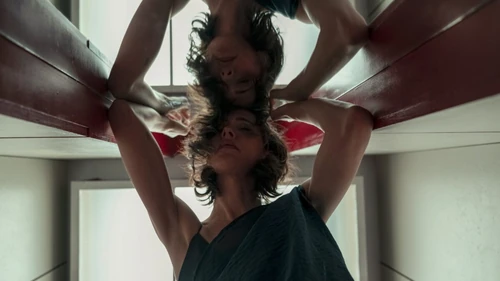 The End of It: «Una mujer sostiene a otra figura sobre sus hombros en una composición simétrica dentro de una habitación.»